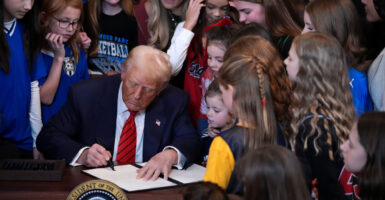 Donald Trump in a suit at a desk signing an executive order, surrounded by women and girls