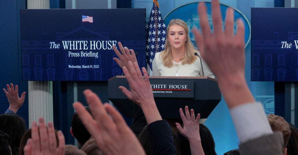 White House press secretary Karoline Leavitt fields questions at a news conference, with reporters raising their hands to be called upon.