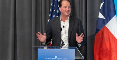 Texas Rep. August Pfluger wears a black suit and white tee shirt while standing at a pulpit.