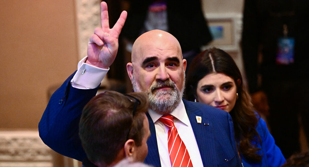 Chris LaCivita in a blue suit with a red tie makes a V for victory symbol