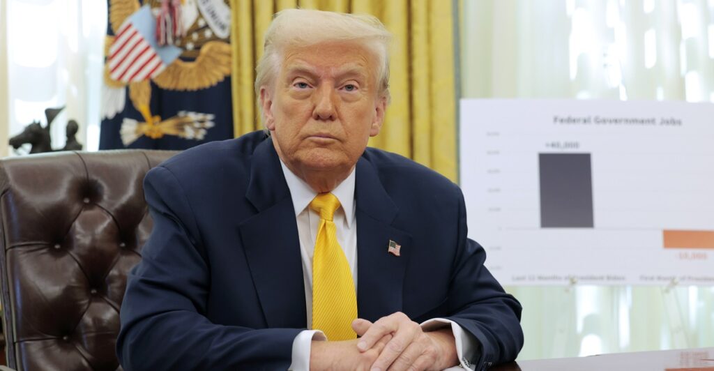 Donald Trump sits at a desk with his hands folded in front of him as he looks straight forward.