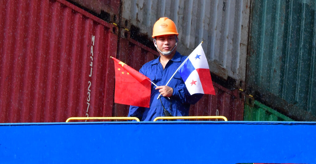 A crew member of a Chinese Cosco Shipping Rose container ship wears a blue jump suit and orange hard hat while holding the Chinese and Panamanian flags.