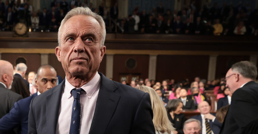 Robert F. Kennedy, Jr. stands wearing a dark suit and dark necktie.