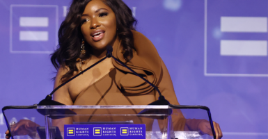Rep. Jasmine Crockett in a light brown dress speaks at a podium in front of a purple background
