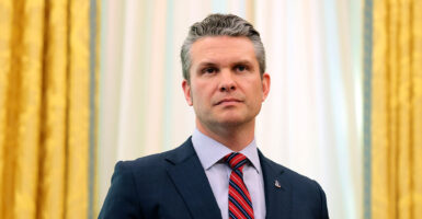 Pete Hegseth stands in front of white and gold curtains while wearing a navy suit.