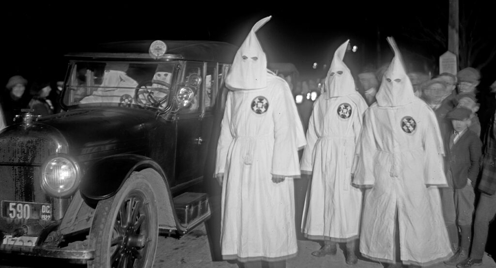 Ku Klux Klan members in white hoods stand next to a black car, driven by Klan members