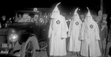 Ku Klux Klan members in white hoods stand next to a black car, driven by Klan members