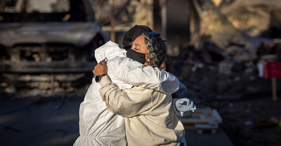 Los Angeles fire victims wearing protective gear embrace among the ashes of their burned homes.