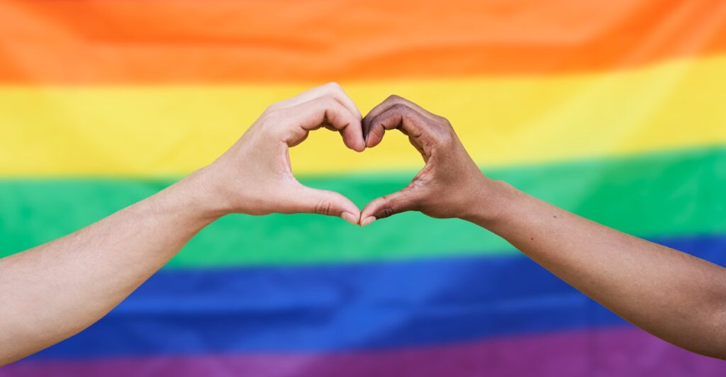 Two hands join together to make the shape of a heart in front of a gay pride flag.