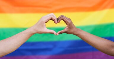 Two hands join together to make the shape of a heart in front of a gay pride flag.