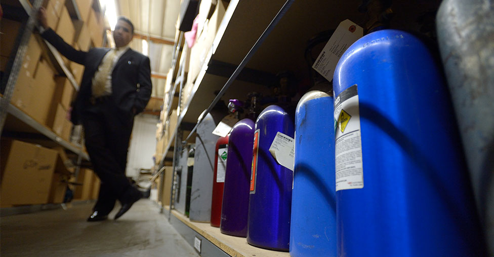 A detective stands beside a shelf of nitrous oxide cannisters in a storage building.