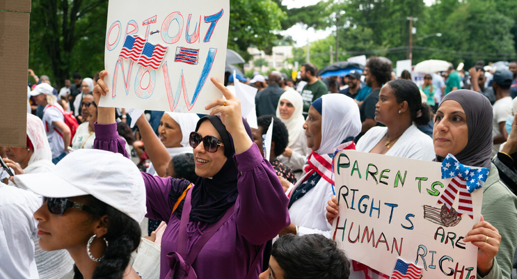 Women wearing head coverings hold signs reading "Opt-Out Now" and "Parents Rights Are Human Rights."