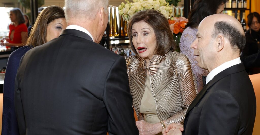 Nancy Pelosi talks with guests who are formed in a circle around her at a party.