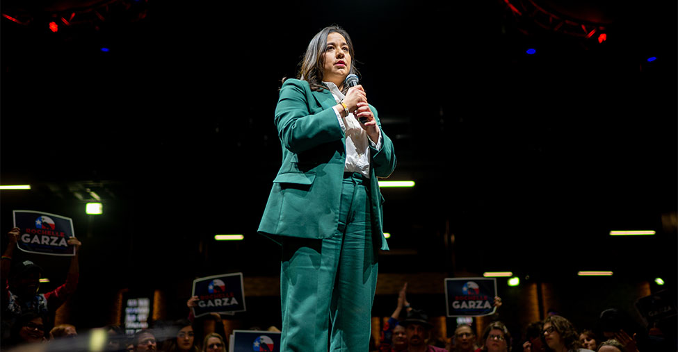 Rochelle Garza holding a microphone while standing on stage wearing a green blazer.