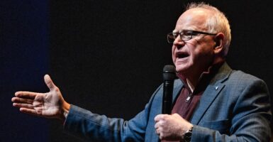 Tim Walz stands with a microphone in his left hand and his right hand reaching out as he speaks to an audience.