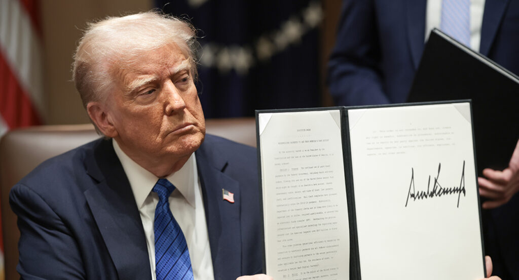 President Donald Trump in a suit with a blue tie holds up an executive order he signed.