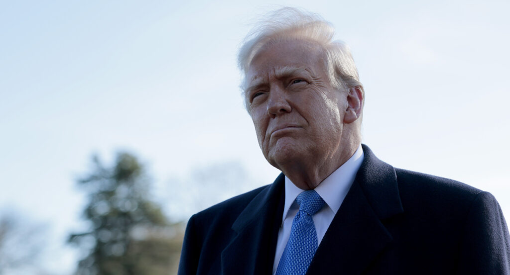 Donald Trump in a black suit with a blue tie looks bemused outside
