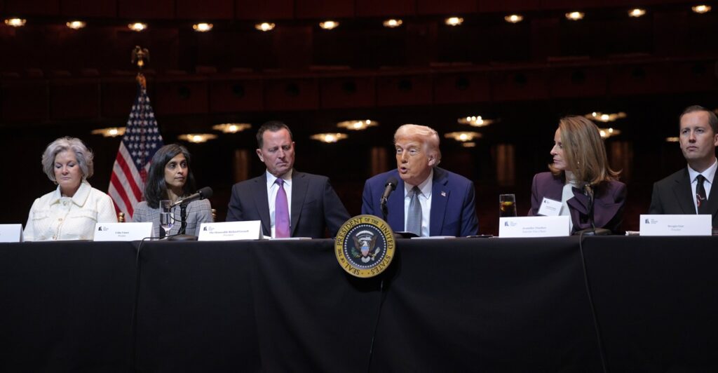 Donald Trump sits at a table and looks out at reporters with both arms at his side.