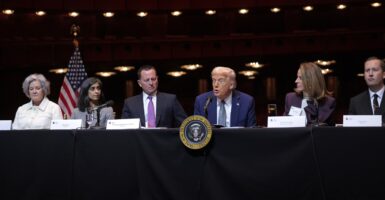 Donald Trump sits at a table and looks out at reporters with both arms at his side.