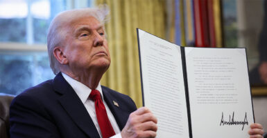 Donald Trump sitting in the oval office wearing a navy suit and red tie while holding up a signed executive order.