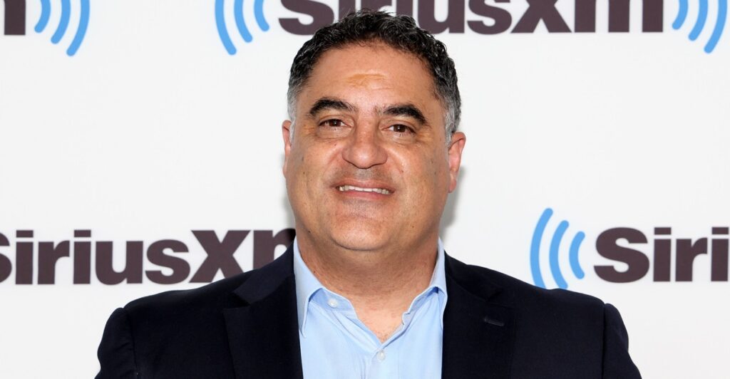Cenk Uygur stands on a red carpet during an event and smiles at the camera.