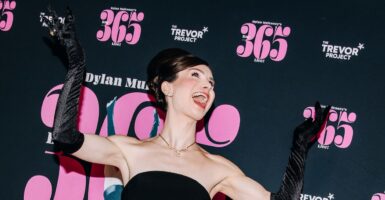 Dylan Mulvaney has both hands raised and is reacting with mouth open while on the red carpet.