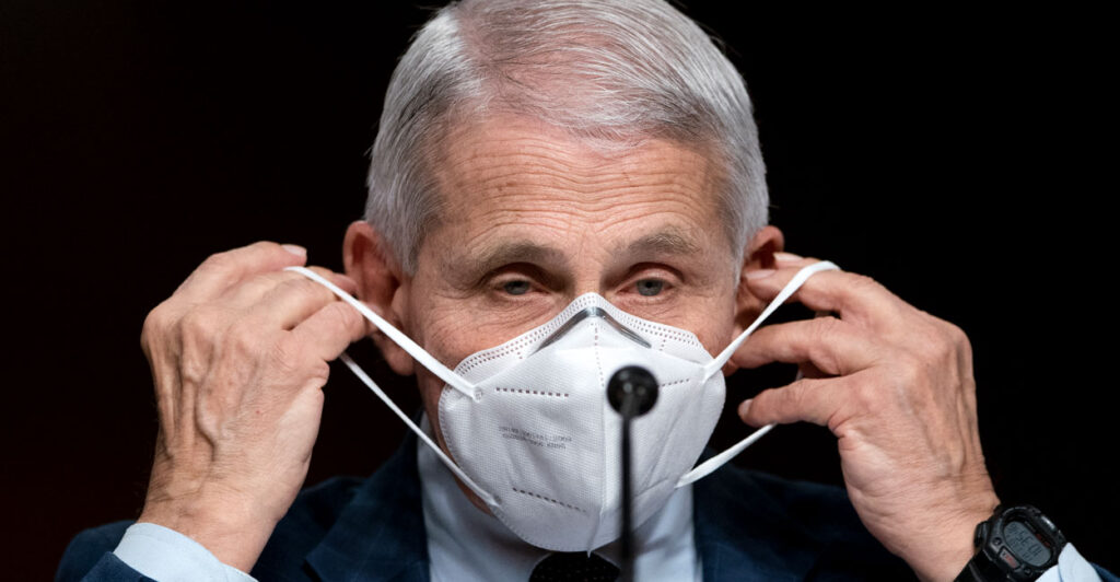 Anthony Fauci in a suit putting on an N95 mask