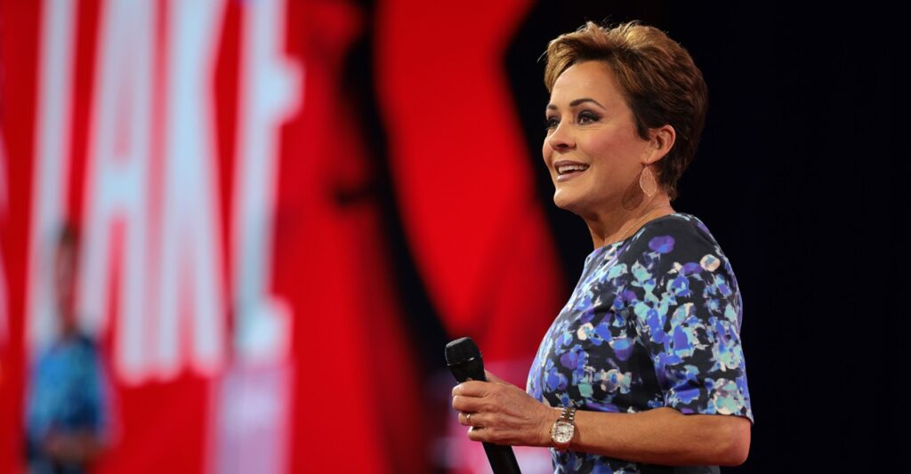 Kari Lake stands and holds a microphone in her left hand and looks out at the audience.