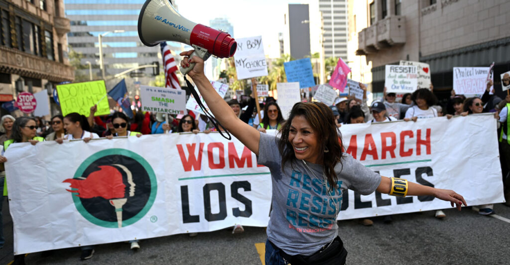 a woman raising her hands with a megaphone leads a protest march through the streets of Los Angeles with women with protest signs and holding a large "Women's March Los Angeles" banner.
