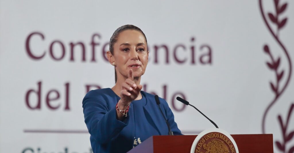 Claudia Sheinbaum Pardo stands in front of a podium and a microphone and holds out her right hand while talking.