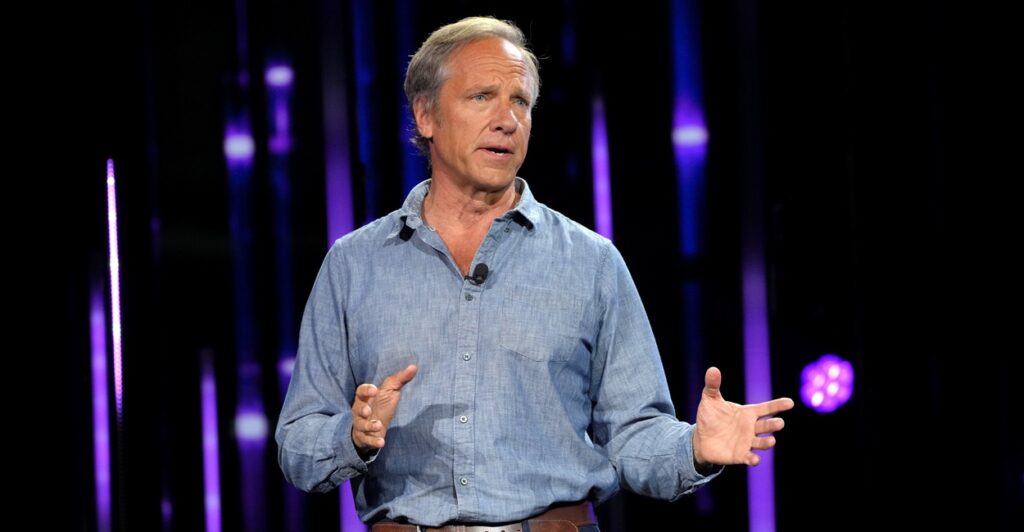 Mike Rowe stands on a stage while talking and holds both hands in front of him to make a point.