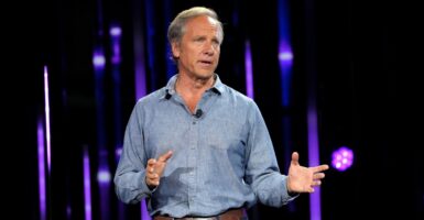 Mike Rowe stands on a stage while talking and holds both hands in front of him to make a point.