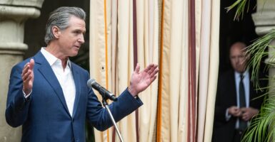 Gavin Newsom stands in front of a microphone with his arms spread out as he speaks to an audience.