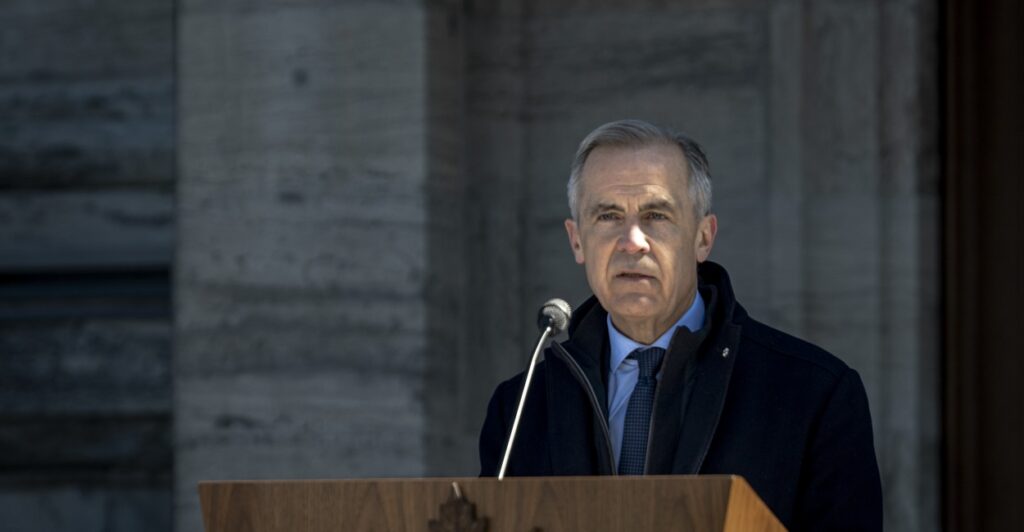 Mark Carney stands at a podium and looks out ahead with a microphone in front of him.