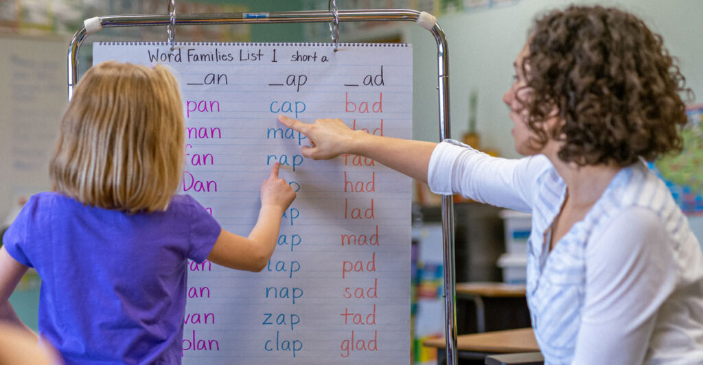 a six-year-old girl in a purple top and her teacher in a white top point to a chart with simple words on it like cat and hat