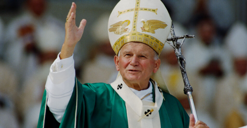 Pope John Paul II with his papal miter and a silver crucifix staff in one hand and raising another hand toward the audience