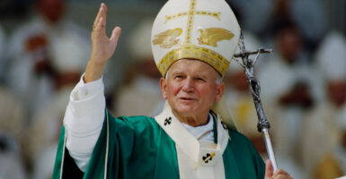 Pope John Paul II with his papal miter and a silver crucifix staff in one hand and raising another hand toward the audience