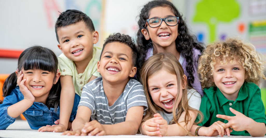 Six cheerful kindergartners laying on classroom floor, smiling at the camera.