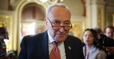 Chuck Schumer in a dark suit and red tie with glasses down his nose looks intently ahead