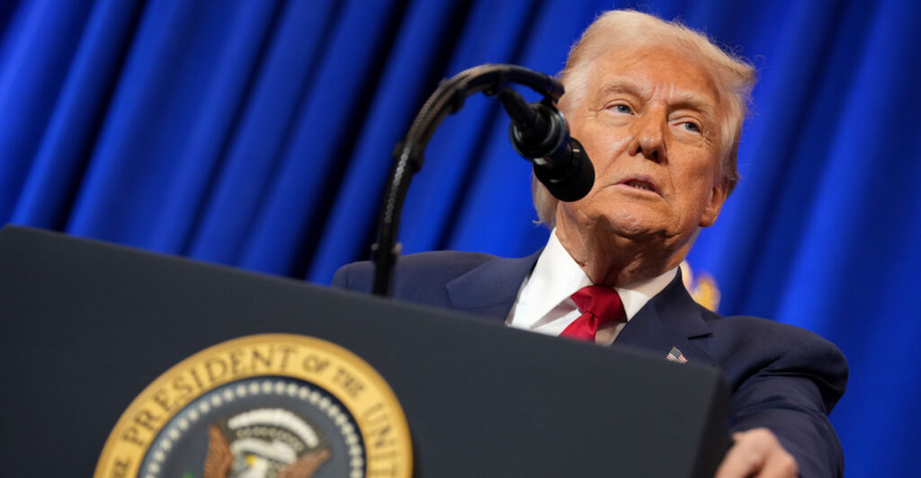Donald Trump in a blue suit and red tie speaking into a microphone at a presidential podium