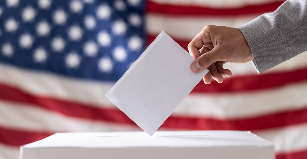 A voter drops his ballot into a ballot box with an American flag unfurled as backdrop.