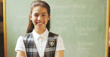 A young girl, wearing private-school attire, stands with arms crossed in front of a chalkboard.