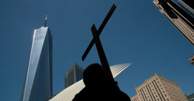 A low-angle shot of a man in silhouette carrying a cross, against a backdrop of the Freedom Tower other high-rises and a perfect blue sky.