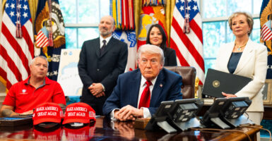 President Donald Trump sitting at his desk in the Oval Office surrounded by his cabinet members and others