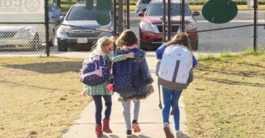 Three little schoolgirls wearing backpacks, about eight-years-old walk toward a gate to leave school. Blonde girl on left has arm draped on back of the middle girl.