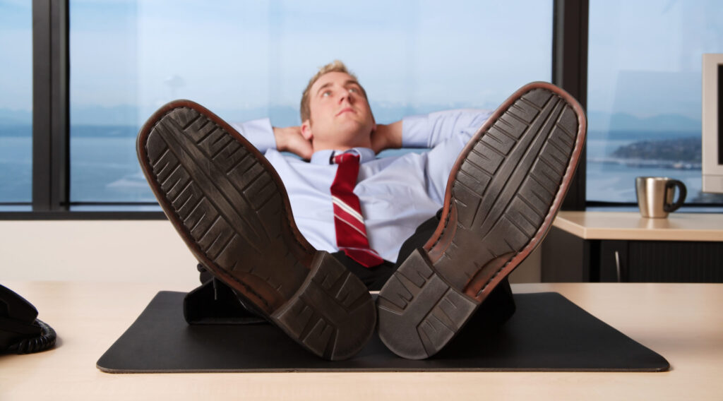 Man in dress shirt and tie leans back in his chair, feet on his desk, hands behind his head, looking up at the sky