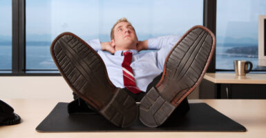 Man in dress shirt and tie leans back in his chair, feet on his desk, hands behind his head, looking up at the sky
