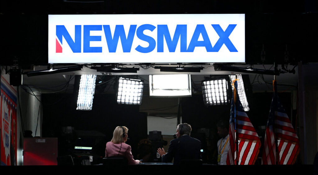 Rear-view shot of two Newsmax news anchors behind a desk underneath stage lights and large blue Newsmax sign on white background.