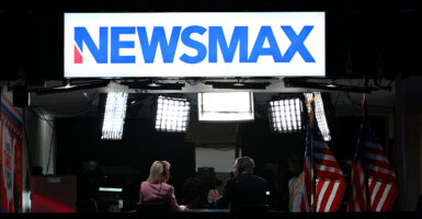 Rear-view shot of two Newsmax news anchors behind a desk underneath stage lights and large blue Newsmax sign on white background.