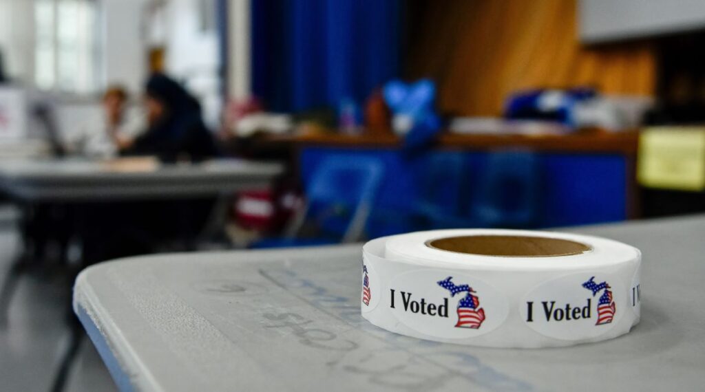 A roll of "I Voted" stickers lays on a grey table, as voting activity takes place out of focus in the background.
